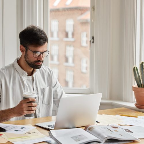 Professional IT developer works on design of new site, studies documentation, holds takeaway coffee. Focused concentrated young graphic designer develops new strategy, poses in cozy interior